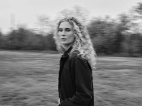 Monochrome portrait of a woman with long curly hair wearing a dark jacket outdoors with motion blur background.