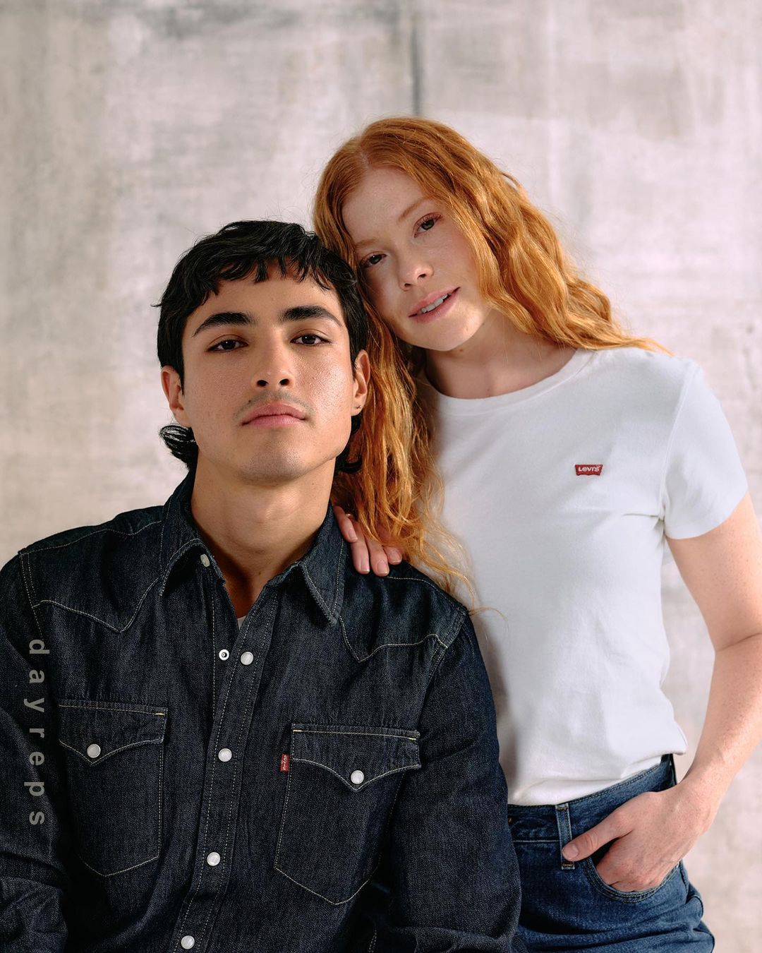 Portrait of a young couple wearing denim in front of a mottled gray backdrop in a studio. The woman is resting her head on the man's head.