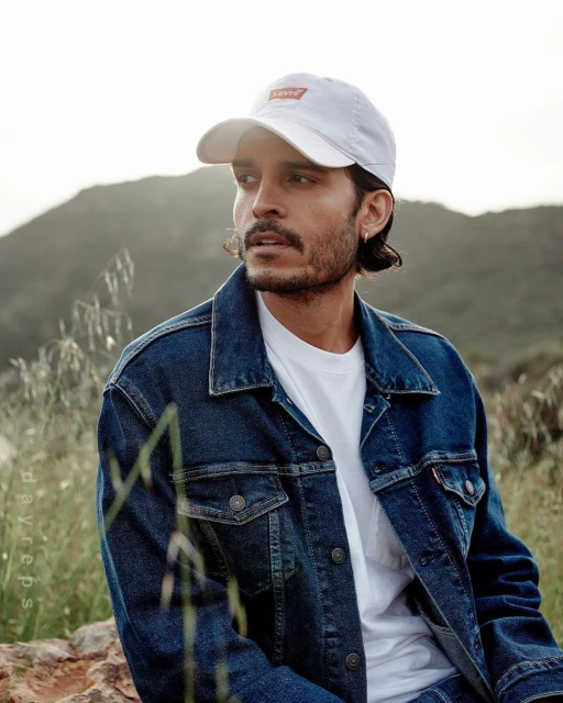 Man in a denim jacket and white baseball cap poses outdoors with blurred mountains in the background, creating a casual, cool vibe.