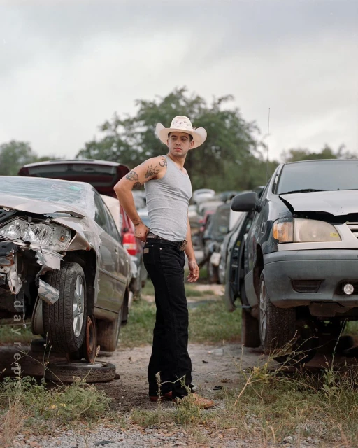 Man in cowboy hat, tank top, and jeans stands in junkyard between two wrecked cars, posing in natural light on an overcast day.