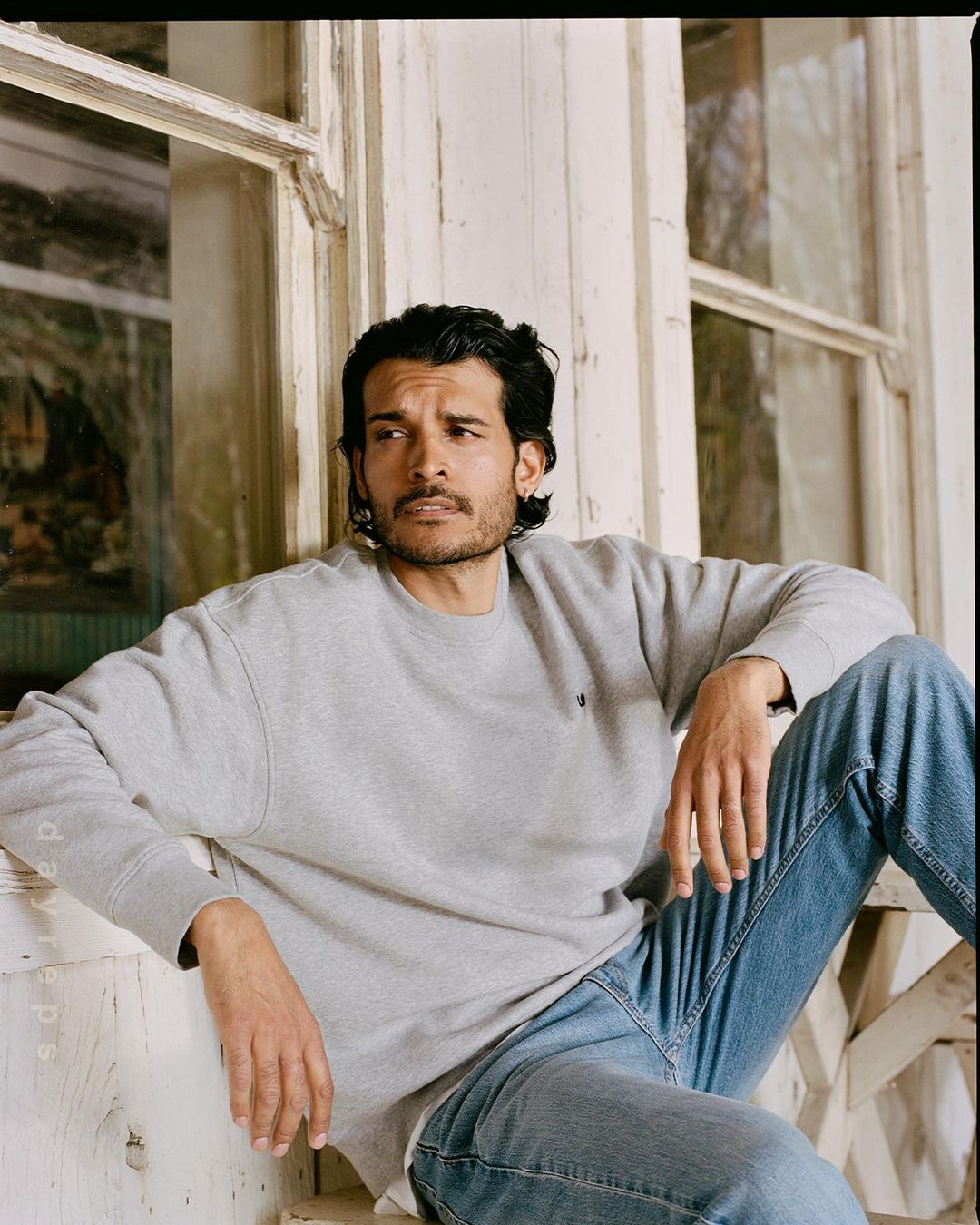Portrait of a man with dark hair wearing a grey sweatshirt and jeans, sitting by a weathered window with a thoughtful expression.