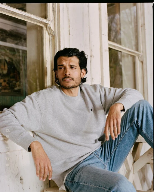 Portrait of a man with dark hair wearing a grey sweatshirt and jeans, sitting by a weathered window with a thoughtful expression.