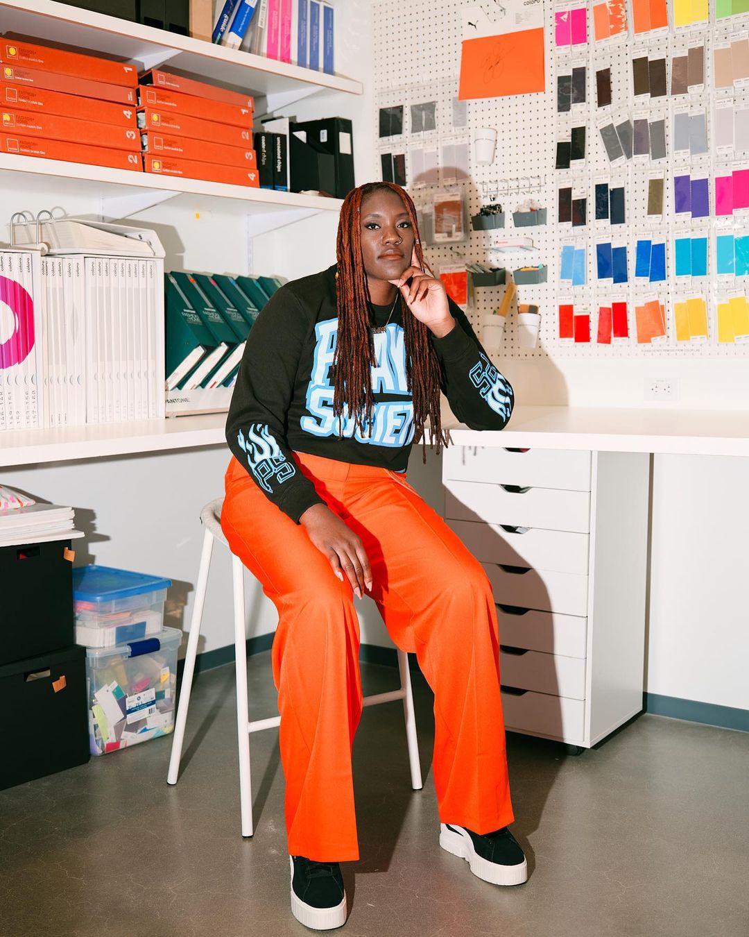 A black woman with braided hair sits on a stool in an artist studio wearing orange pants and a black printed sweatshirt with white shelves.