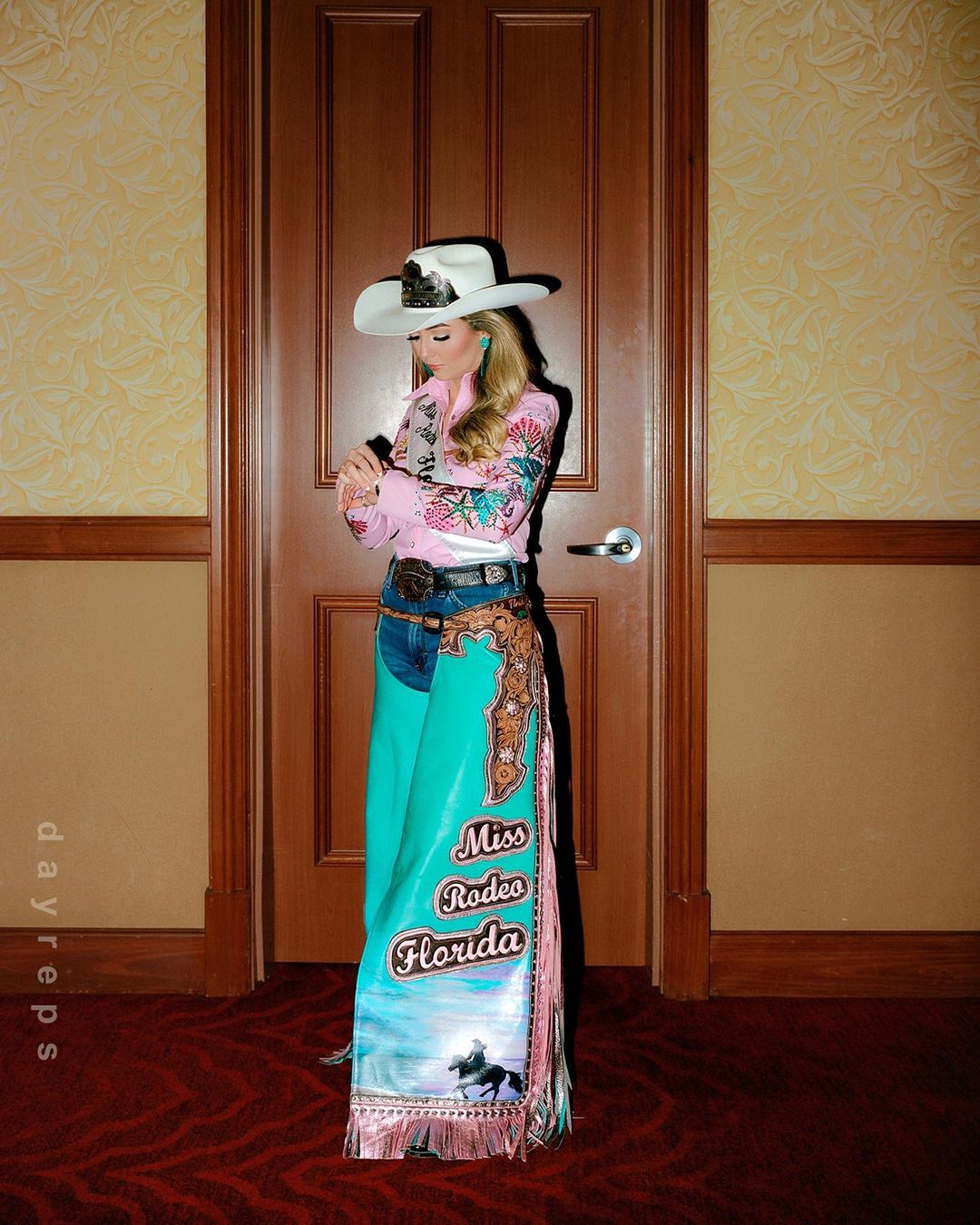 A woman wearing a cowgirl hat and rodeo attire, standing in front of a closed door with floral wallpaper and red carpet.