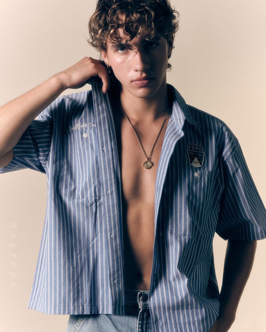 Portrait of a young adult male model with curly hair, wearing an unbuttoned blue-and-white striped shirt and a pendant necklace.