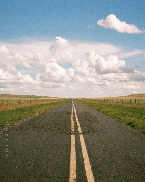 Straight asphalt road leads through a grassy field on a clear day with scattered clouds in the sky, creating a sense of open travel.