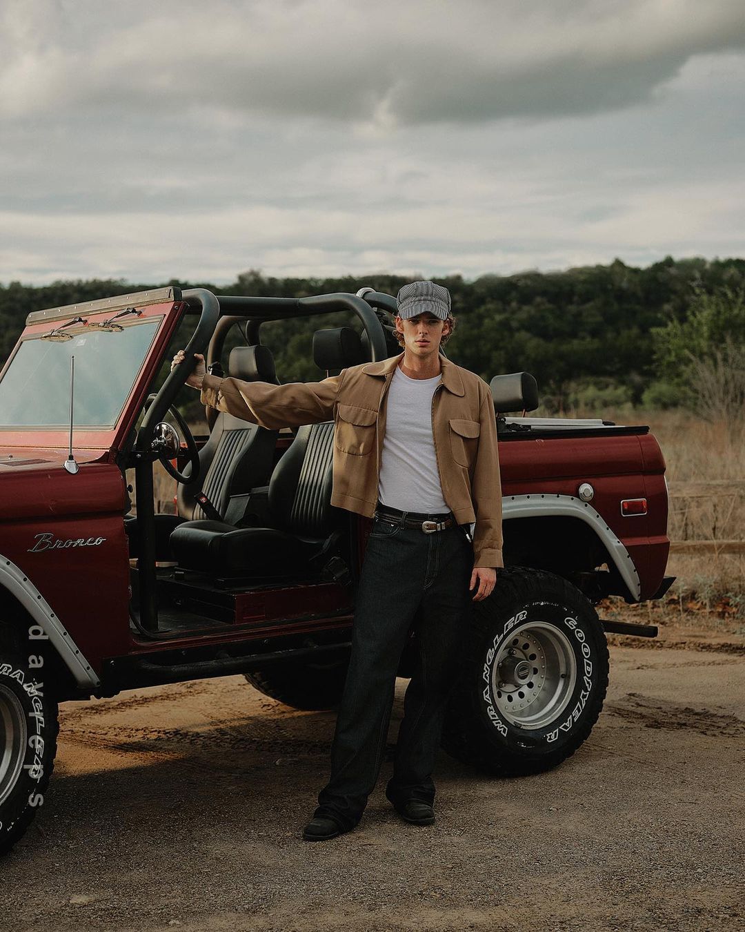 A young man poses next to a red Bronco in an outdoor setting. He wears a cap, jacket, t-shirt, and jeans in a casual pose.