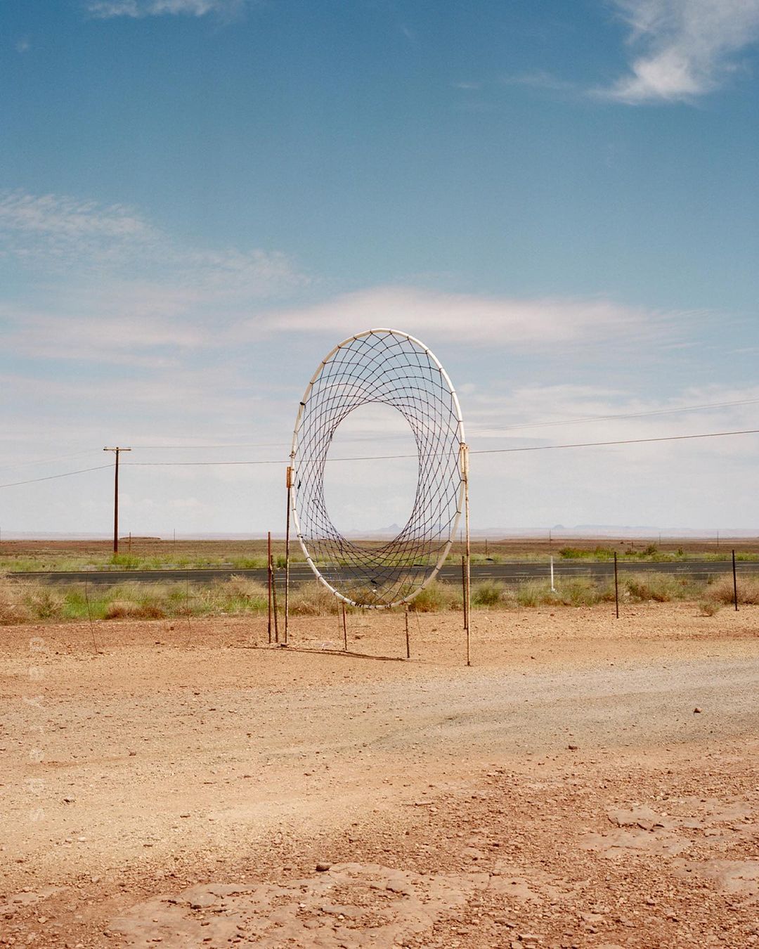 Open-air sculpture of a large circular net structure in a desert landscape under a blue sky with scattered clouds on a sunny day.