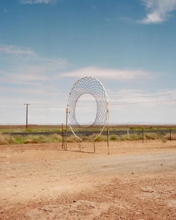 Open-air sculpture of a large circular net structure in a desert landscape under a blue sky with scattered clouds on a sunny day.