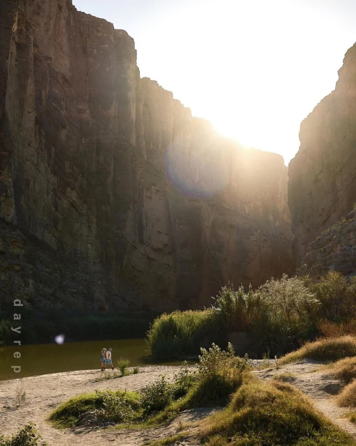 Scenic canyon landscape with bright sunlight, sandy shoreline, tall reeds, and two people on the edge of calm water in the distance.