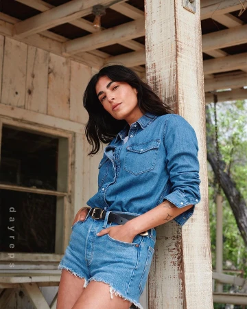 Woman with dark hair wearing denim shirt and shorts leaning against a wooden porch pillar outdoors.