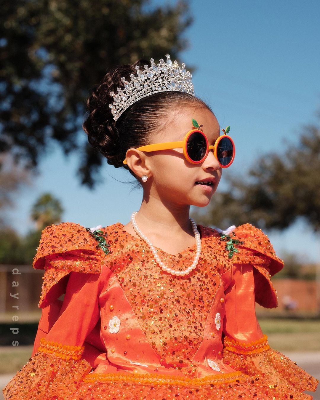 Young child wearing an orange sequined dress, sparkling tiara, round orange sunglasses, and pearl necklace against an outdoor background.