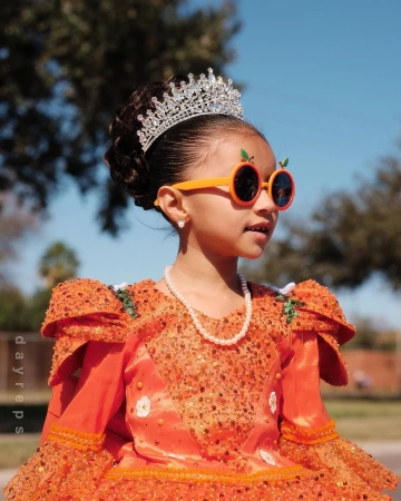 Young child wearing an orange sequined dress, sparkling tiara, round orange sunglasses, and pearl necklace against an outdoor background.