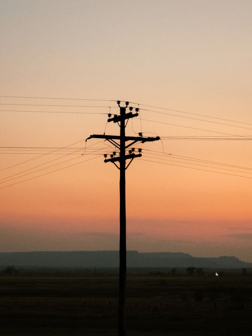 Silhouette of a utility pole at sunset. Electrical power lines extend across the frame against a colorful gradient sky.