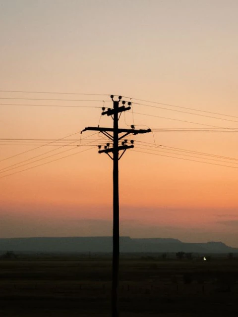 Silhouette of a utility pole at sunset. Electrical power lines extend across the frame against a colorful gradient sky.