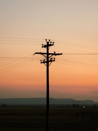Silhouette of a utility pole at sunset. Electrical power lines extend across the frame against a colorful gradient sky.