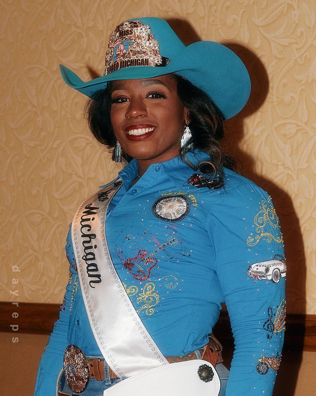 Portrait of a smiling African-American woman wearing a cowboy hat and Miss Michigan sash, standing indoors in front of a wall.