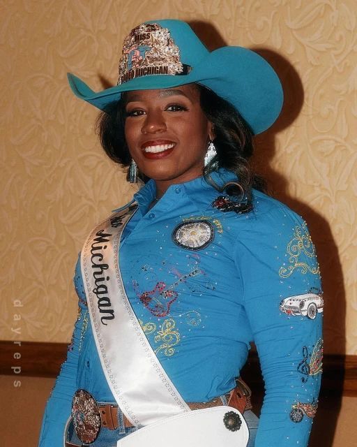 Portrait of a smiling African-American woman wearing a cowboy hat and Miss Michigan sash, standing indoors in front of a wall.