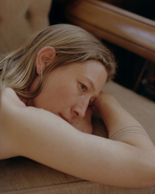 Blonde woman lies on a tan couch looking left with arms raised; light skin and a tattoo on the left arm are visible.
