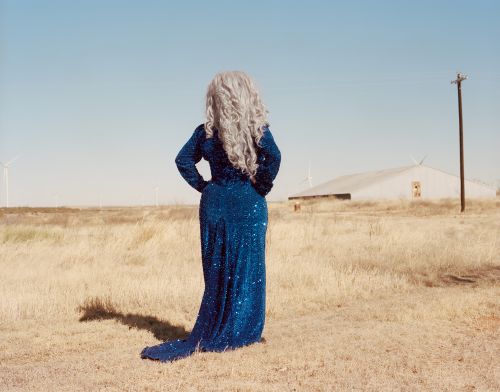 A person in a long, shimmering blue dress stands in a sunlit, grassy field with a building and wind turbines in the distance.