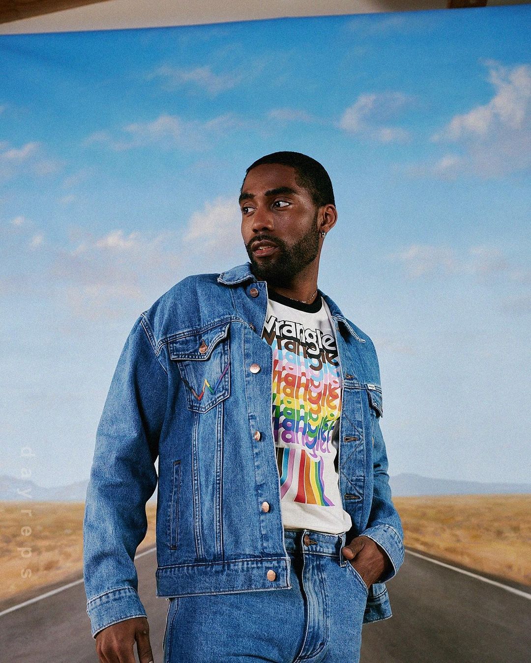 Adult male in a denim jacket and jeans stands on a road against a sky backdrop, showcasing fashion and casual style.