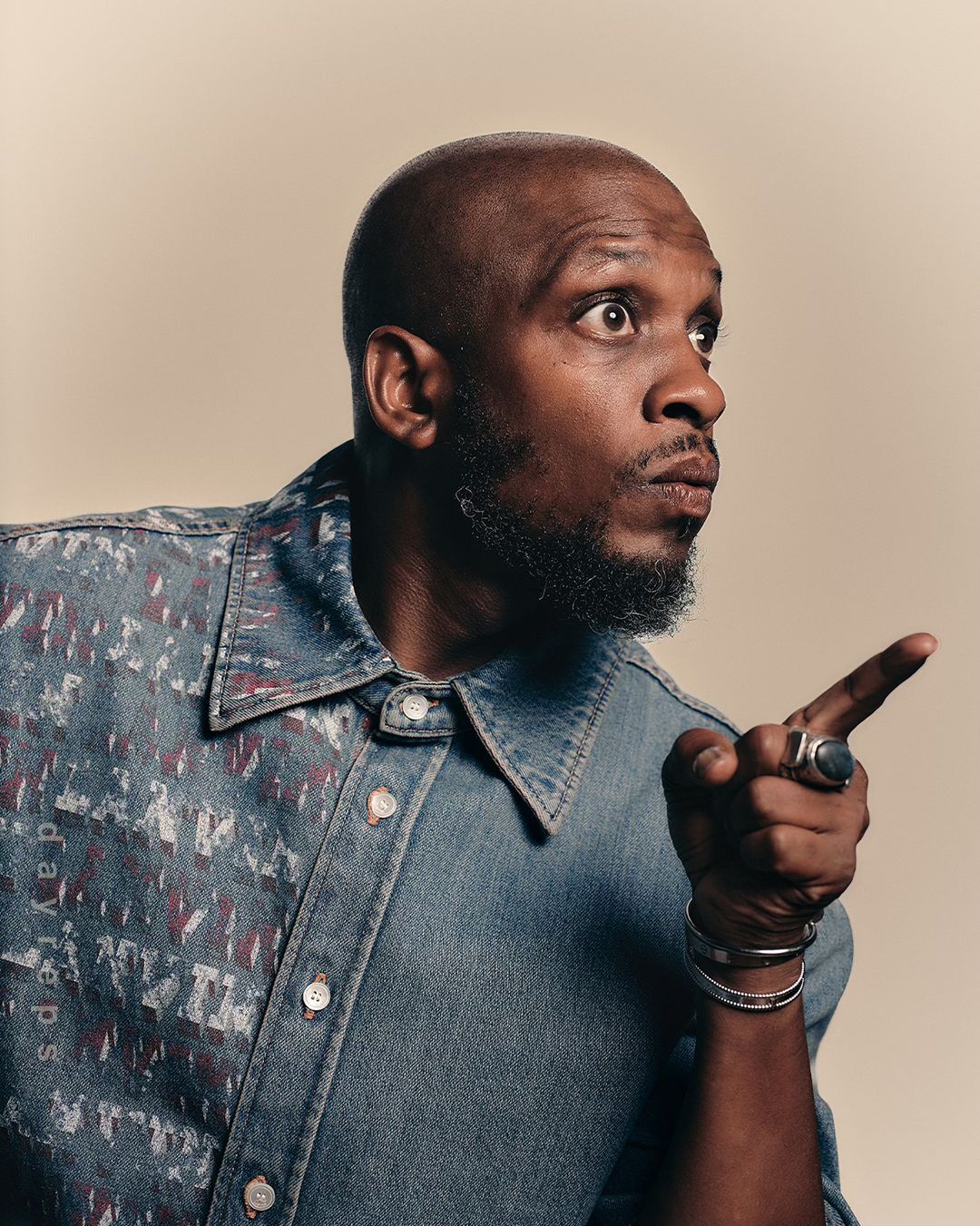 Close-up shot of a stylish bald man pointing to the side with a surprised expression wearing a unique denim button-down shirt.