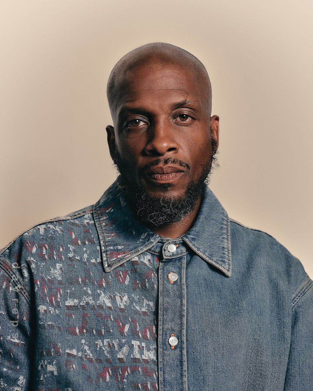 Close-up portrait of a bald, bearded African-American adult male wearing a color-blocked denim shirt against a neutral backdrop.