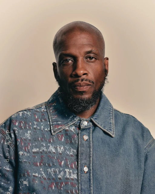 Close-up portrait of a bald, bearded African-American adult male wearing a color-blocked denim shirt against a neutral backdrop.