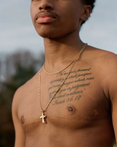 Young man with chest tattoo and gold necklaces posing outdoors against blurred background. He appears shirtless in natural daylight.