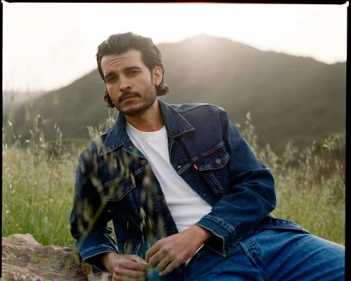 An adult male with dark hair and a moustache sits outside in a denim jacket and jeans with a white t-shirt in a natural setting.