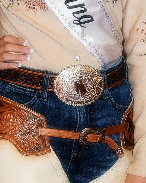 Midsection of a person wearing blue jeans, a tan western shirt, a rodeo belt buckle, and cowboy chaps, showcasing western fashion.