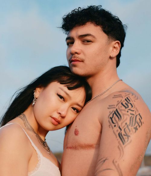 An Asian woman rests her head on a Caucasian man's shoulder against a blue sky backdrop, both adorned with silver necklaces.