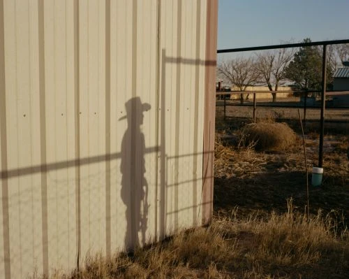 Outdoor shot featuring a shadow of a person in a cowboy hat on a corrugated metal wall, set against a backdrop of dry grassy terrain.