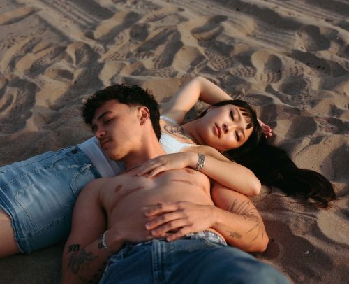 A shirtless man and an Asian woman lie intertwined on a sandy beach. The couple embraces as the sun sets, casting warm tones.