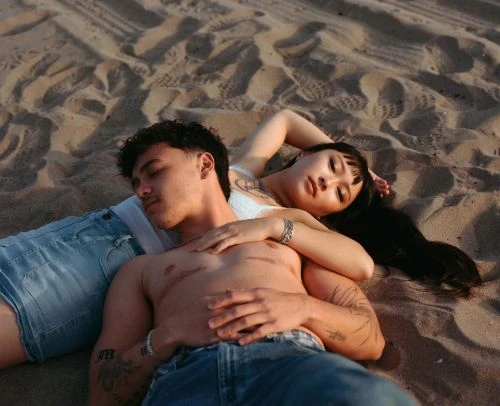 A shirtless man and an Asian woman lie intertwined on a sandy beach. The couple embraces as the sun sets, casting warm tones.