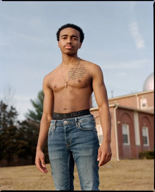 Young man poses outdoors shirtless in jeans with visible Fruit of the Loom waistband, set against a blurred sky and building backdrop.