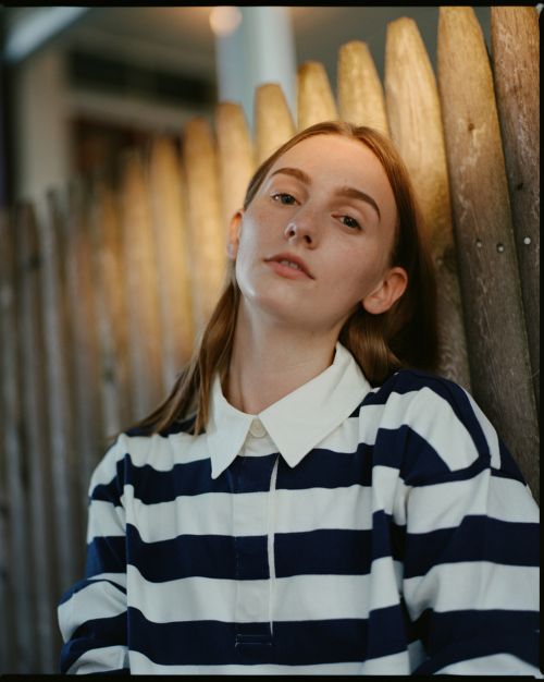 Close-up shot of a young adult female model wearing a striped top, leaning against a weathered wooden fence, looking towards the camera.