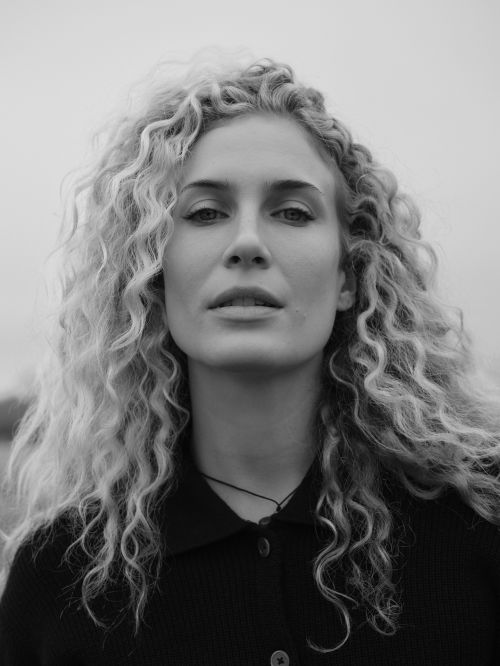 Black and white portrait of a young woman with curly blond hair, wearing a collared shirt against an outdoor background.