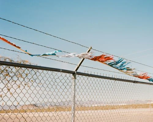 Weathered red, white, and blue streamers hang on a chain-link fence against a clear blue sky, adding a festive yet distressed touch to the scene.