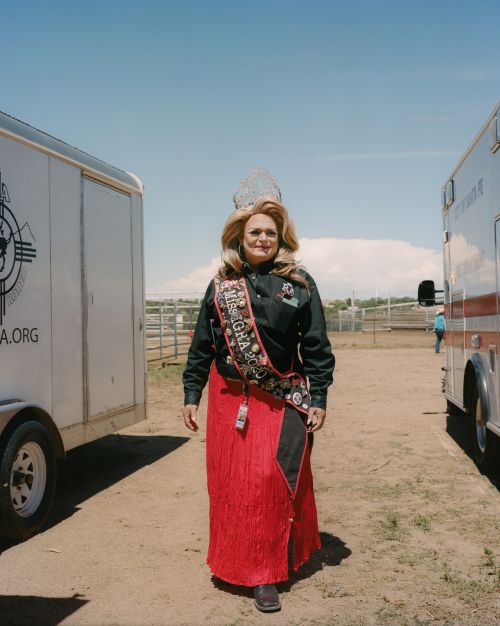 A woman wearing a crown and 'Miss IGRA' sash stands between trailers and a fence on a dirt lot under a sunny sky.