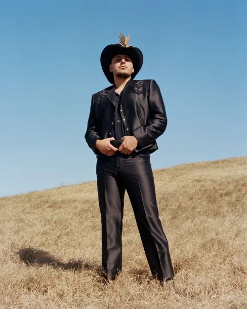 Adult male wearing a cowboy hat and black jacket in a golden field under a blue sky poses with hands near belt buckle.
