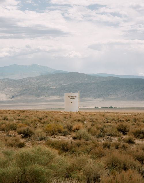 A white cylindrical tank stands in a field of desert scrub with mountains and sky visible in the background on a cloudy day.