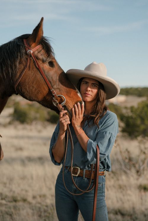 Adult female in a cowboy hat stands next to a brown horse in a rural outdoor setting, under a clear sky. The woman is holding the horse's face.