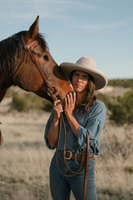 Adult female in a cowboy hat stands next to a brown horse in a rural outdoor setting, under a clear sky. The woman is holding the horse's face.