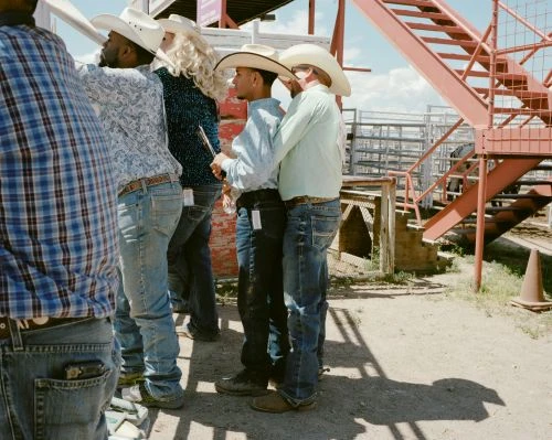 Group of people wearing cowboy hats and western attire standing outdoors with a metal structure and light sky in the background.