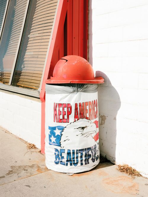 Patriotic trash can topped with an orange hard hat near a white brick building with red trim and a window on a sunny day.