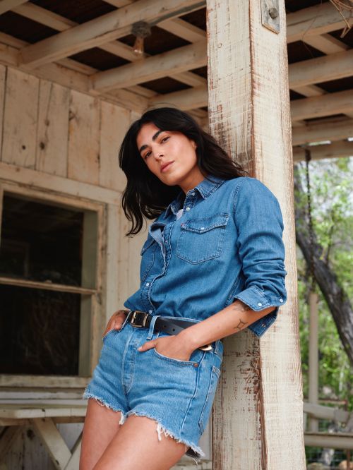 A woman in a denim shirt and shorts poses outdoors, leaning against a wood post with a window and green foliage in the background.