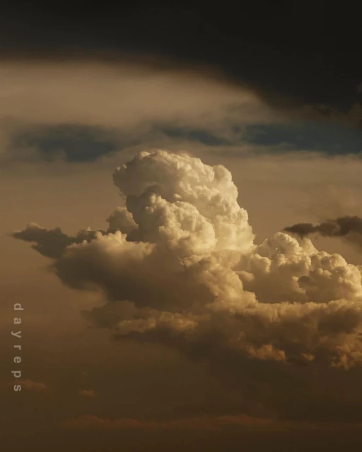 Dramatic cumulus clouds fill the frame in this low-angle shot, displaying a range of gold and gray tones against a darkened sky.