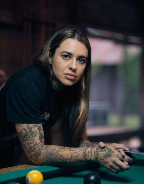 Portrait of an adult woman with tattoos, leaning on a pool table with billiard balls, indoors with natural light, medium shot, serious expression.