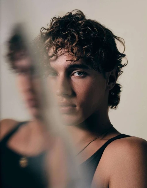 Portrait of a young adult male with curly brown hair and blue eyes, wearing a black tank top, against a neutral background with diffused lighting.